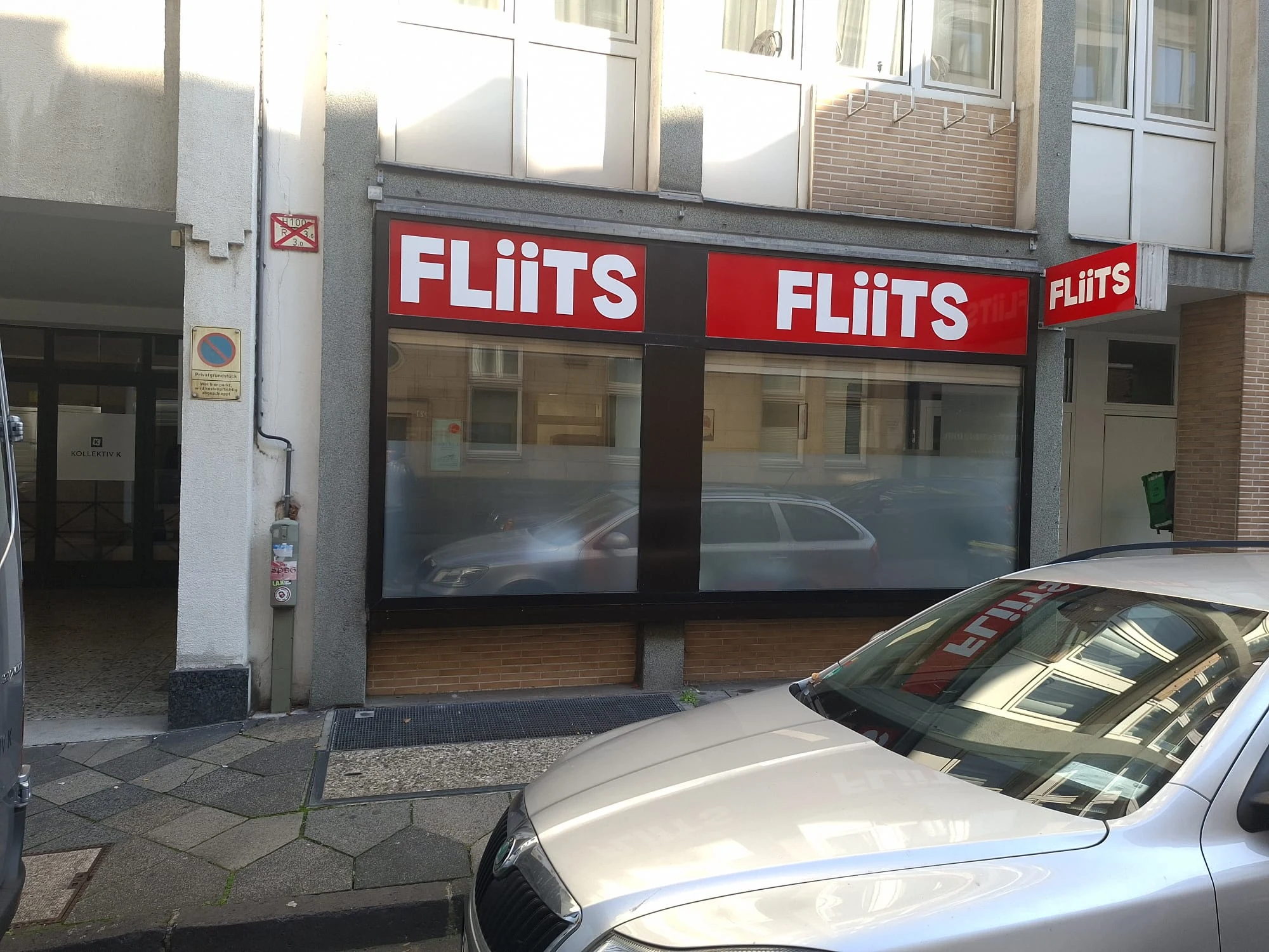 Fassade von FLiiTS in der Alexanderstraße 22 in Düsseldorf: rote Blenden mit weißem FLiiTS-Logo über beiden Schaufenstern, daneben der beschriftete Leuchtkasten