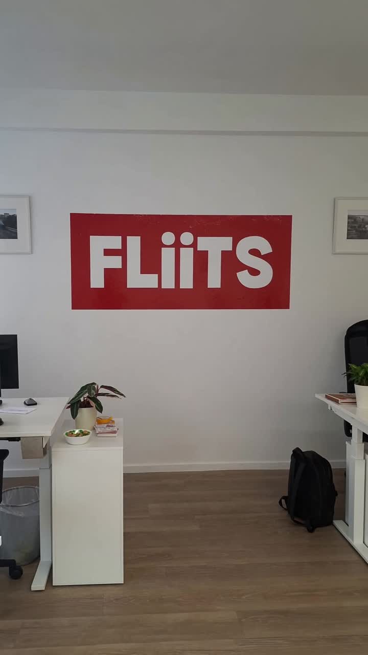 KI-rekonstruiertes Standbild des leeren FLiiTS-Büros: weiße Wortmarke auf roter Fläche an der Innenwand